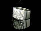 Close-up image of a white gold ring encrusted with multiple small diamonds, reflecting light on a black surface. The ring features a pattern of closely set diamonds creating a sparkling effect.